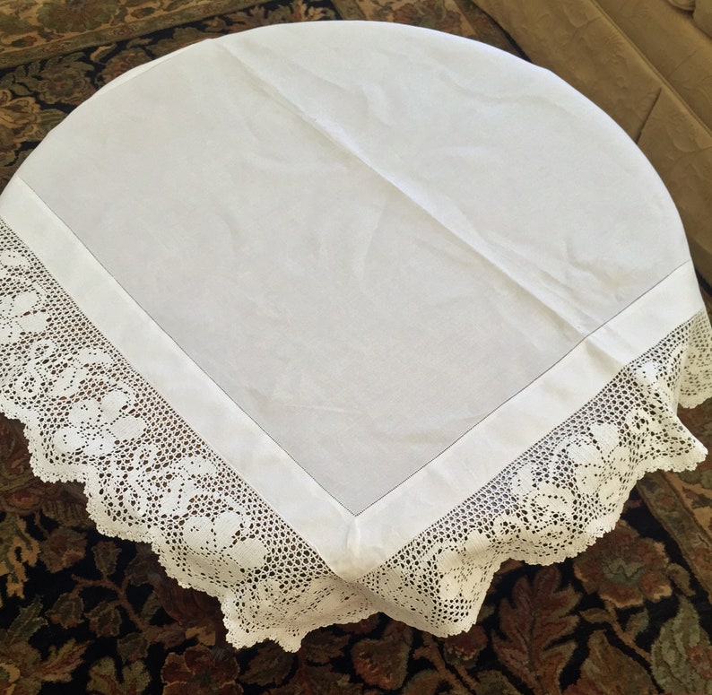 May include: White round tablecloth with a delicate white crocheted lace border. The tablecloth is made of a smooth, linen-like fabric.