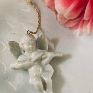May include: A white porcelain angel ornament with wings and a gold string for hanging. The angel is playing a musical instrument.