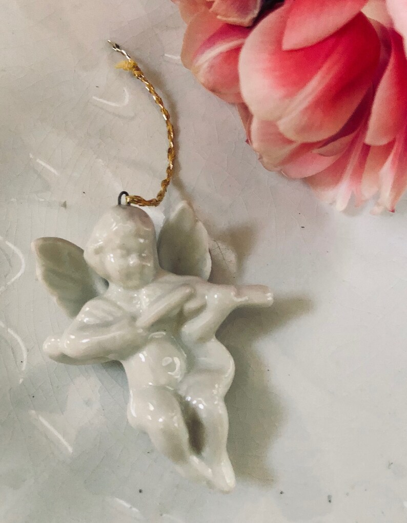 May include: A white porcelain angel ornament with wings and a gold string for hanging. The angel is playing a musical instrument.