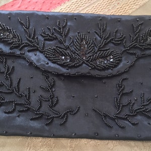Puede incluir: Un bolso de mano de sat&eacute;n negro con un dise&ntilde;o floral hecho de cuentas negras. El bolso tiene un cierre de solapa y un ribete de cuentas.