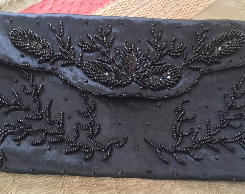 Puede incluir: Un bolso de mano de sat&eacute;n negro con un dise&ntilde;o floral hecho de cuentas negras. El bolso tiene un cierre de solapa y un ribete de cuentas.