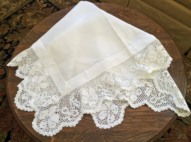 May include: White linen tablecloth with a delicate crocheted lace border. The lace features a floral design.