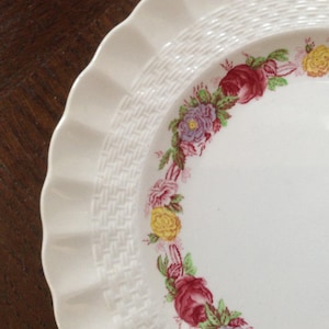 May include: White ceramic plate with a woven pattern and a floral border. The flowers are pink, yellow, purple, and green.