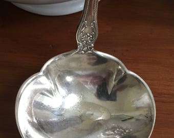 May include: A silver ladle with an ornate handle and a rounded bowl. The ladle is polished and reflects light.