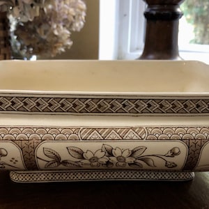 May include: A white ceramic serving dish with a brown floral pattern. The dish has a rectangular shape with rounded edges and a raised base.