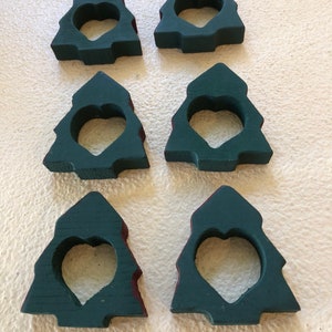 May include: Six green wooden Christmas tree napkin rings with a heart-shaped cutout. The rings are painted a dark green with a red accent around the edges.