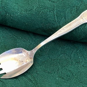 May include: A silver salad serving fork with a spoon on the other end. The handle is decorated with an ornate design.