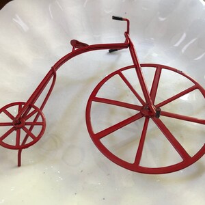 May include: A red metal toy bicycle with a large front wheel and a smaller back wheel. The bicycle is made of thin metal and has a simple design.