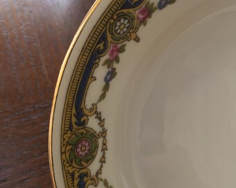 May include: A white ceramic bowl with a gold rim and a blue and gold floral design. The bowl has a scalloped edge and is decorated with pink, blue, and yellow flowers.