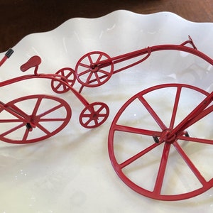 May include: A red metal sculpture of a bicycle with a large front wheel and a smaller back wheel. The bicycle is on a white plate.