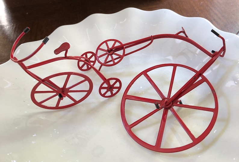 May include: A red metal sculpture of a bicycle with a large front wheel and a smaller back wheel. The bicycle is on a white plate.