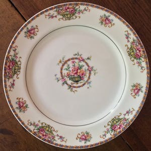 May include: A white ceramic dinner plate with a gold and blue trim. The plate features a floral design with pink, yellow, and green flowers.