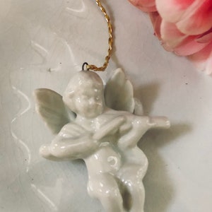 May include: A white porcelain angel ornament with wings, holding a musical instrument. The ornament has a gold string attached to the top for hanging.