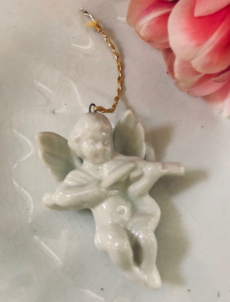 May include: A white porcelain angel ornament with wings, holding a musical instrument. The ornament has a gold string attached to the top for hanging.