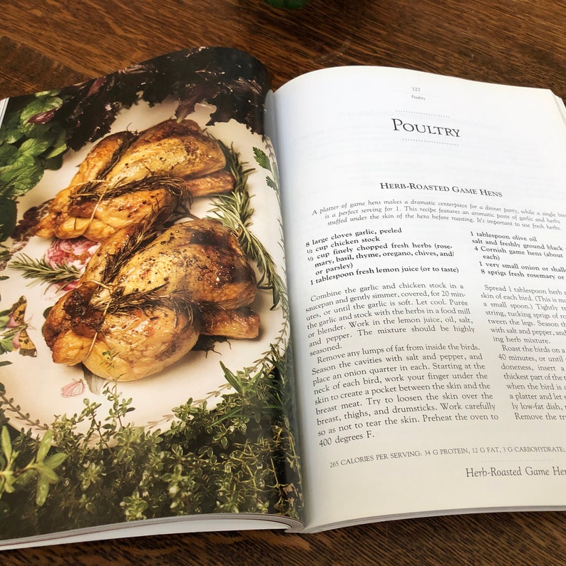 May include: A close-up of a cookbook page with a recipe for Herb-Roasted Game Hens. The recipe includes ingredients like garlic, fresh herbs, chicken stock, and lemon juice. The page also includes a photo of two roasted game hens on a white plate with rosemary sprigs.
