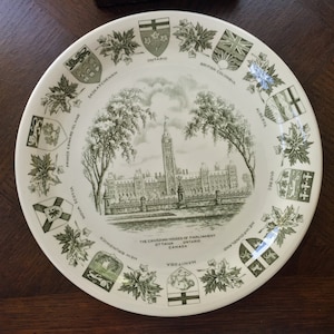 Puede incluir: Un plato de cerámica blanco con un diseño verde y blanco que representa las Casas del Parlamento canadiense en Ottawa, Ontario, Canadá. El plato está decorado con los escudos de armas de cada provincia y territorio canadiense.