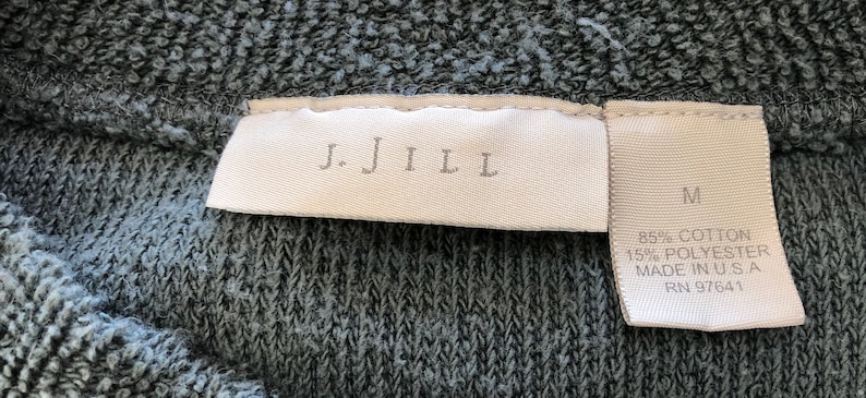 May include: A white tag with the text "J.Jill" and a white tag with the text "M 85% Cotton 15% Polyester Made in U.S.A. RN 97641" on a dark gray fabric.