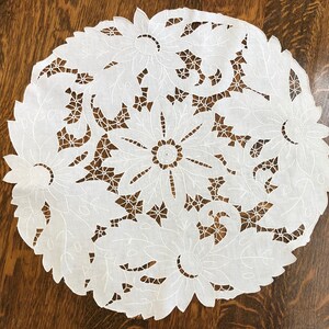 May include: White cotton doily with a delicate floral design. The doily is round and features intricate cutouts of flowers and leaves.