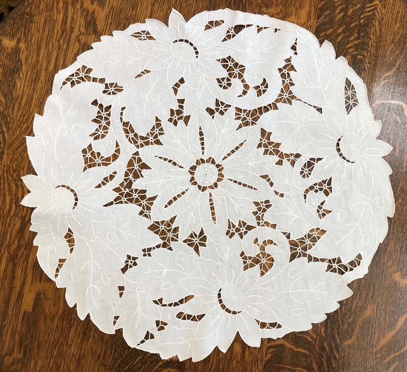 May include: White cotton doily with a delicate floral design. The doily is round and features intricate cutouts of flowers and leaves.