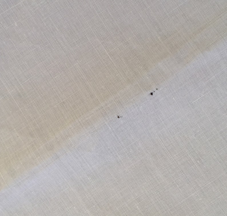 May include: A close-up of a white fabric surface with a subtle texture. There are two small dark spots on the fabric.