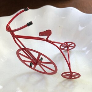 May include: A red metal toy bicycle with a single large front wheel and two smaller back wheels. The bicycle has a curved handlebar and a small seat.