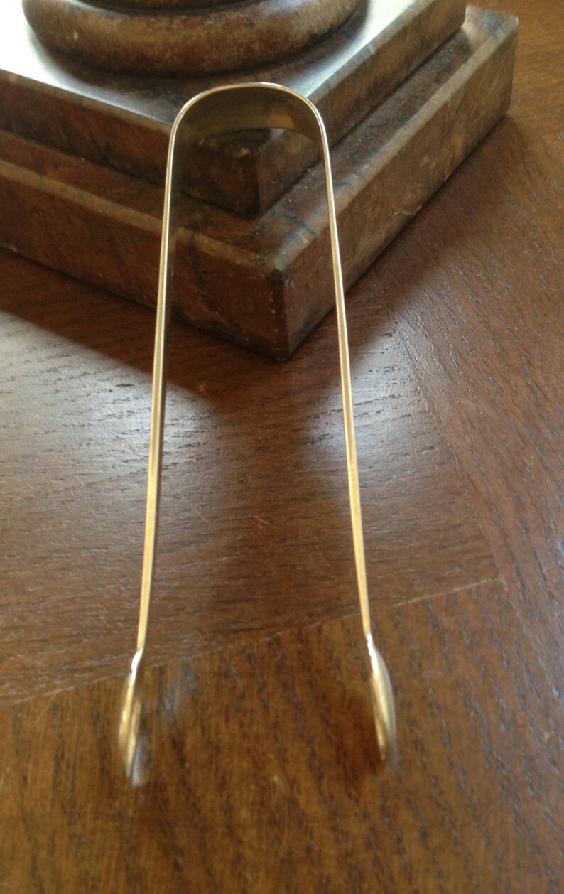 May include: Silver sugar tongs with rounded ends. The tongs are lying on a wooden surface.