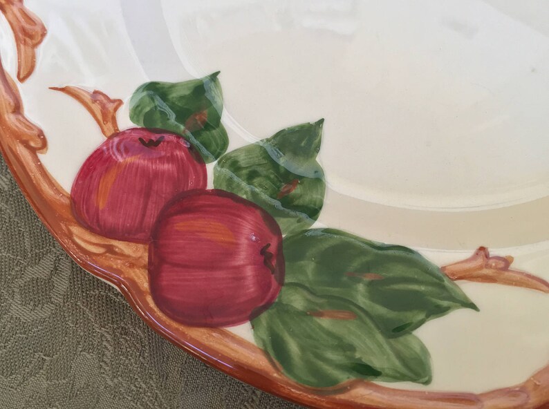 Pu&ograve; includere: Un piatto in ceramica bianca con un bordo marrone e un disegno dipinto di mele rosse e foglie verdi.