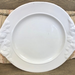 May include: A white ceramic serving platter with two ornate handles. The handles are sculpted with raised floral designs.