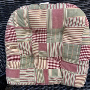 May include: A patchwork chair cushion with a red, green, and cream color scheme. The cushion is made of a soft, quilted fabric and has a square shape.
