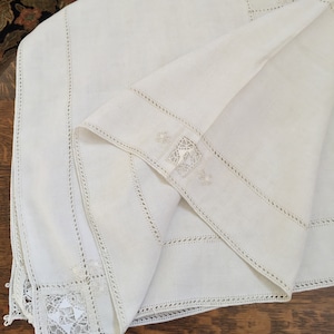 May include: A white linen tablecloth with delicate embroidered lace trim. The tablecloth features a square design with a floral motif in the center.