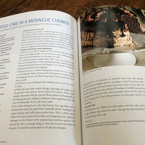 May include: A close-up of a chocolate fudge cake with a meringue topping, topped with a chocolate glaze and pecan halves. The cake is on a white cake stand. The recipe for the cake is visible in the background.