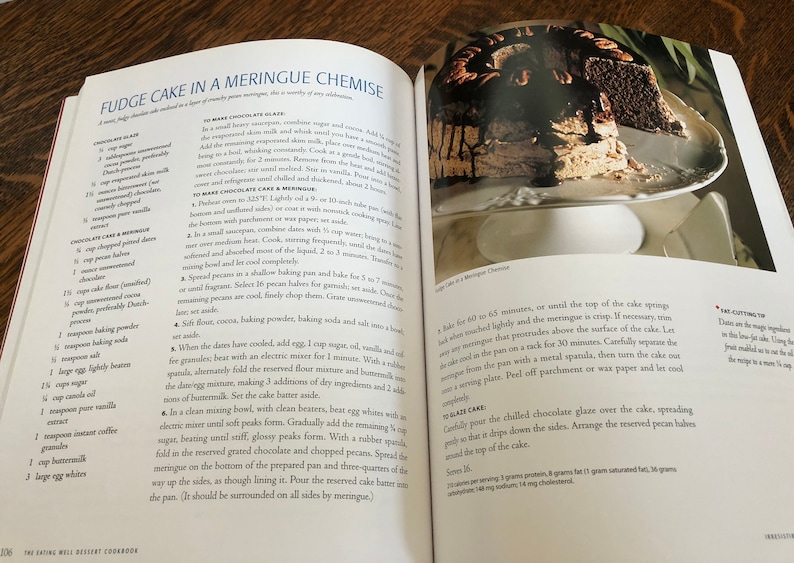 May include: A close-up of a chocolate fudge cake with a meringue topping, topped with a chocolate glaze and pecan halves. The cake is on a white cake stand. The recipe for the cake is visible in the background.