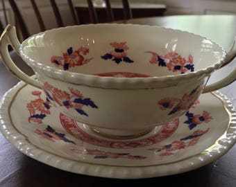 May include: A white porcelain soup bowl with a floral pattern in shades of red, blue, and gold. The bowl has two handles and sits on a matching saucer.