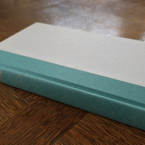 May include: A white hardcover book with a teal blue band around the spine. The title of the book is "The Catcher in the Rye" by J.D. Salinger.