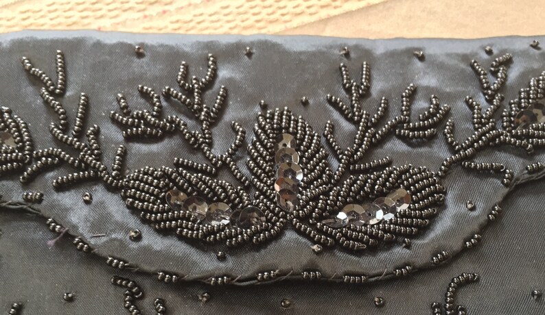 Puede incluir: Un bolso de mano de sat&eacute;n negro con un dise&ntilde;o de cuentas. El dise&ntilde;o presenta cuentas negras y lentejuelas dispuestas en un patr&oacute;n floral.