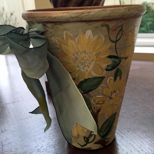May include: A terracotta flower pot with a distressed white finish, painted with yellow flowers and green leaves. A light green ribbon is tied around the pot.