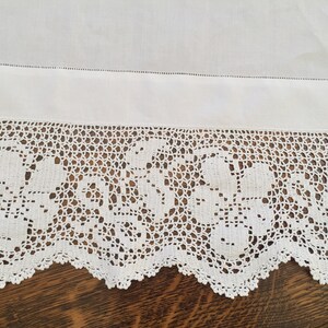 May include: White cotton tablecloth with a crocheted lace border featuring a floral design. The lace is delicate and intricate, with a scalloped edge.