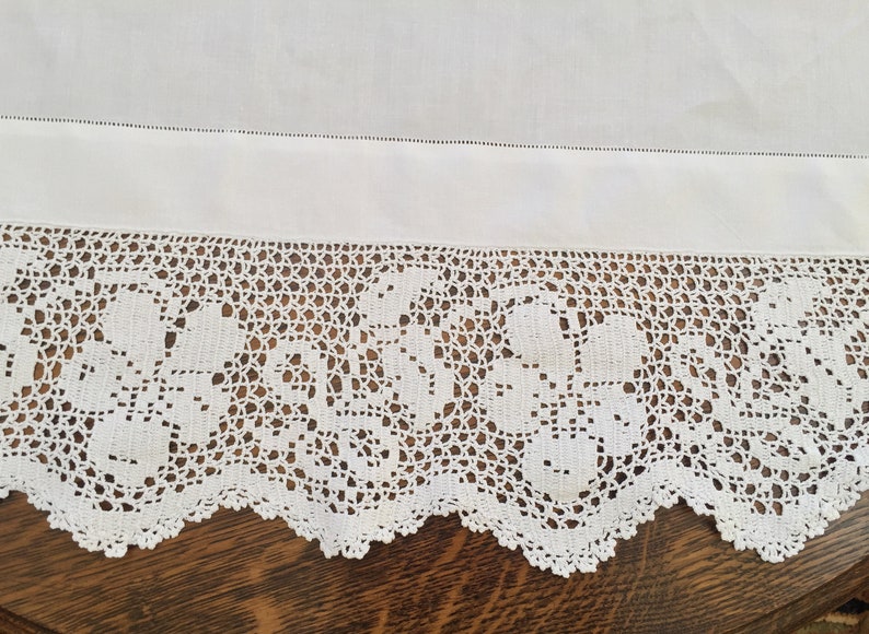 May include: White cotton tablecloth with a crocheted lace border featuring a floral design. The lace is delicate and intricate, with a scalloped edge.