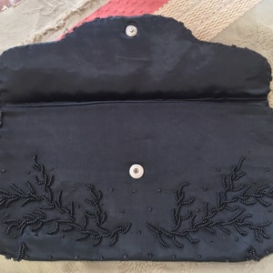 Puede incluir: Un bolso de mano de sat&eacute;n negro con un dise&ntilde;o floral de cuentas. El bolso tiene un cierre de presi&oacute;n y un peque&ntilde;o bolsillo en el interior.