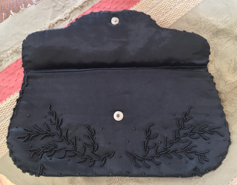 Puede incluir: Un bolso de mano de sat&eacute;n negro con un dise&ntilde;o floral de cuentas. El bolso tiene un cierre de presi&oacute;n y un peque&ntilde;o bolsillo en el interior.