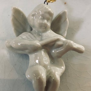 May include: A white porcelain angel figurine with wings, playing a musical instrument. The angel is hanging from a gold chain.