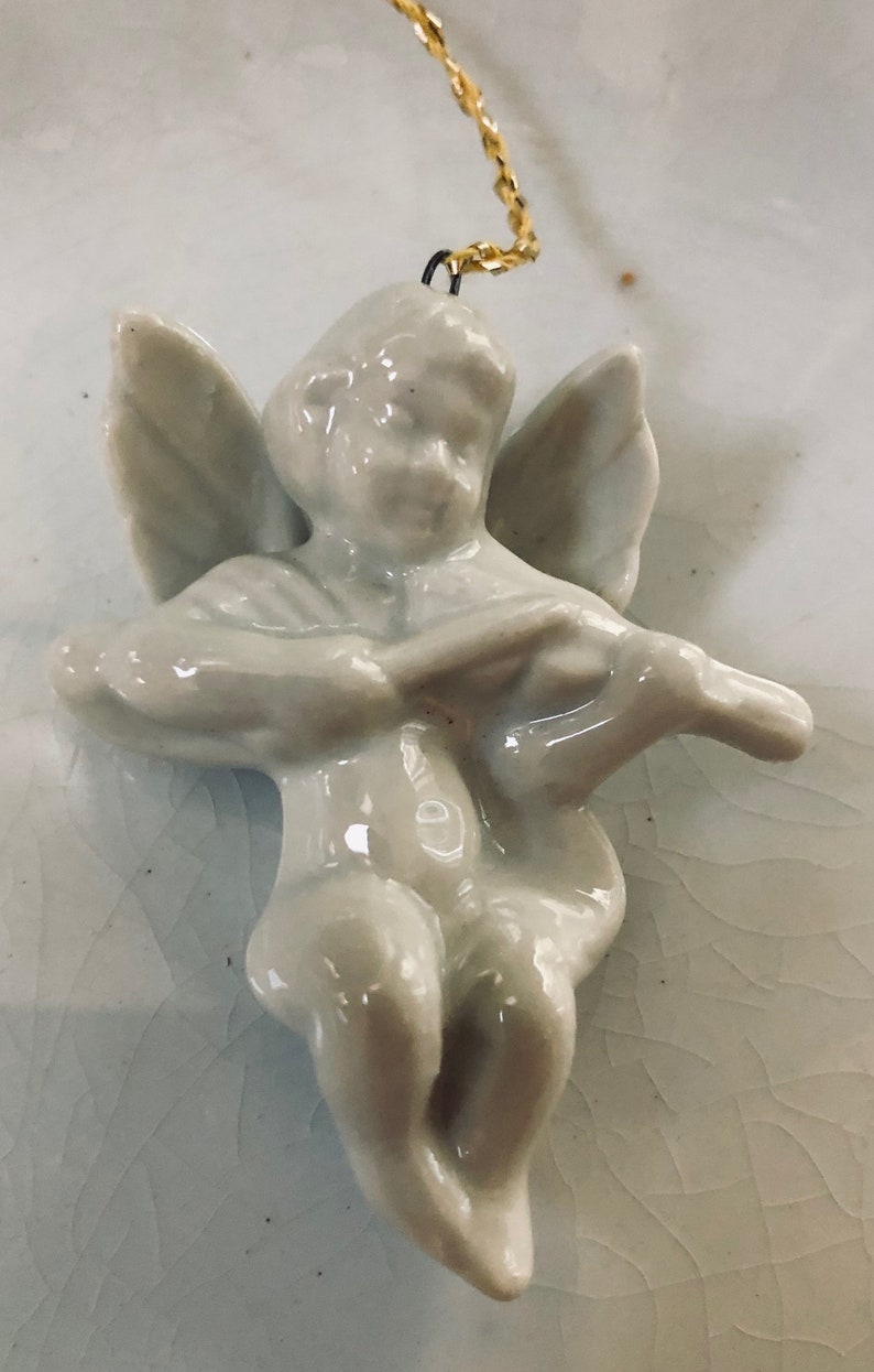 May include: A white porcelain angel figurine with wings, playing a musical instrument. The angel is hanging from a gold chain.
