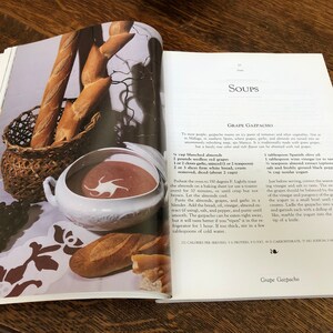 May include: A close-up of a bowl of cold grape gazpacho soup, a recipe from a vintage cookbook. The soup is a vibrant red color and is garnished with a swirl of white yogurt. The cookbook page also includes a list of ingredients and instructions for making the soup.