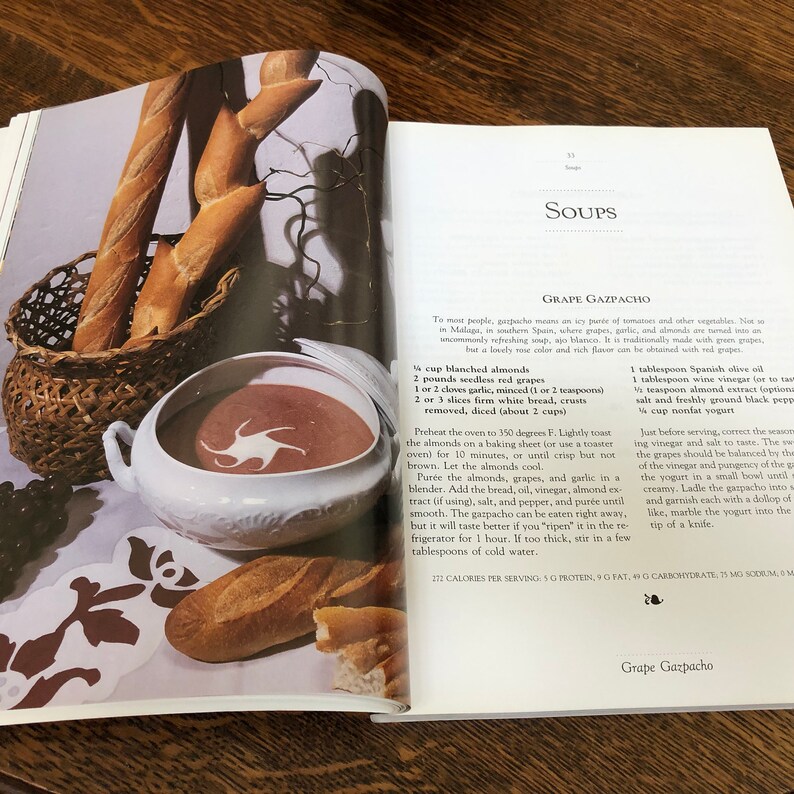 May include: A close-up of a bowl of cold grape gazpacho soup, a recipe from a vintage cookbook. The soup is a vibrant red color and is garnished with a swirl of white yogurt. The cookbook page also includes a list of ingredients and instructions for making the soup.