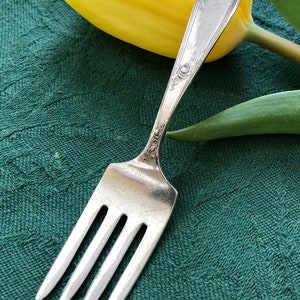 May include: A silver fork with an ornate handle, resting on a green cloth with a yellow tulip behind it.