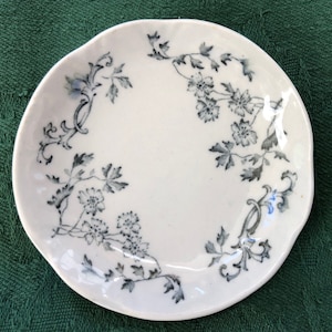 May include: A white ceramic saucer with a scalloped edge and a black and white floral design.