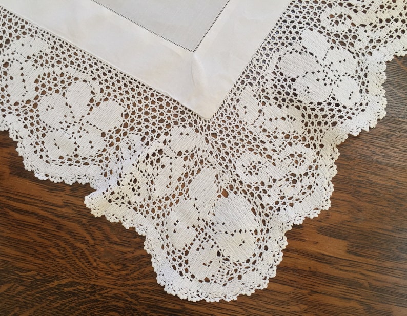 May include: White crocheted tablecloth with a delicate floral pattern. The tablecloth has a scalloped edge and is made of fine, intricate lace.