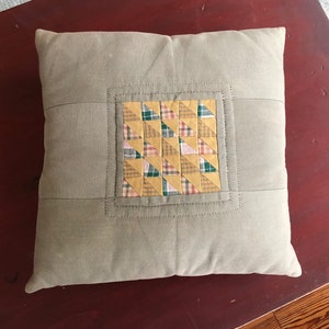 May include: A beige pillow with a square patchwork design in the center. The patchwork is made of yellow, green, pink, and red fabric. The pillow is on a wooden surface.