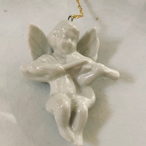 May include: A white porcelain angel ornament with wings, playing a musical instrument. The ornament is attached to a gold string.