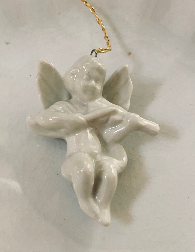 May include: A white porcelain angel ornament with wings, playing a musical instrument. The ornament is attached to a gold string.